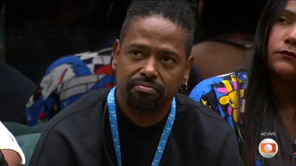 Leandro Boneco, um homem negro de barba e cabelos curtos, observa algo lateralmente com uma expressão de desânimo ou reflexão, usando uma camiseta preta Ao fundo, outras pessoas estão presentes em um ambiente interno, enquanto um logotipo de transmissão ao vivo aparece no canto inferior direito.