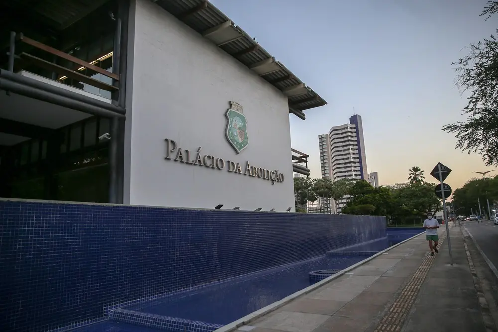 Na imagem, fotografia em ângulo diagonal do Palácio da Abolição, sede do governo do Ceará, em Fortaleza. O edifício possui arquitetura moderna com uma grande parede branca central onde se lê, em letras metálicas, 