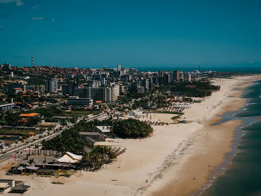 Foto aérea da Praia do Futuro. A imagem mostra parte do mar, faixa de areia com barracas e árvores e diversos prédios que ocupam o bairro.
