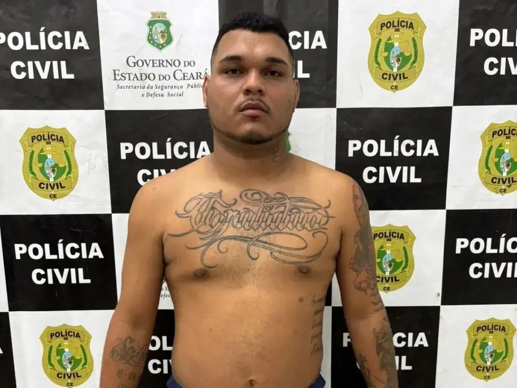 Homem, de frente para a câmera, sem camisa, diante de um painel com os dizeres “polícia civil” e brasões do governo do Ceará. Há tatuagens visíveis no peito e nos braços. Ele é Lucas Lima de Almeida, o Coiote, líder do Comando Vermelho preso no Ceará.
