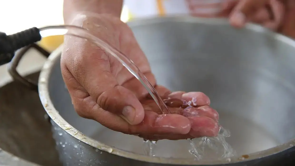 Imagem de uma torneira jorrnado agua em uma bacia. em destaque uma mão se molhando no jato de agua.
