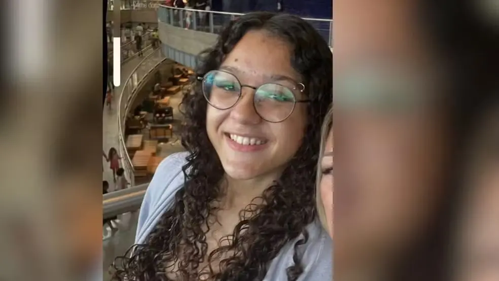 Jovem sorrindo usando óculos e cabelo cacheado, posando para a foto em um ambiente de shopping com várias lojas ao fundo.