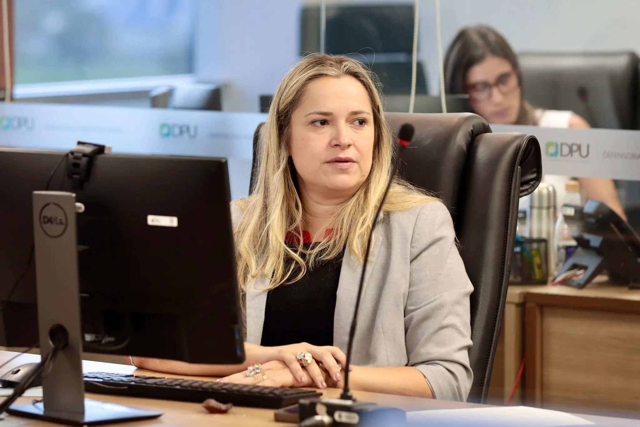 Tarcijany Linhares sentada à mesa durante reunião da DPU. Ela está diante de uma mesa com um computador e um microfone.