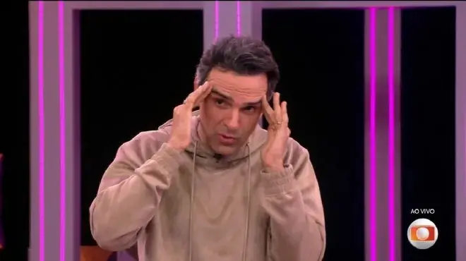 Tadeu Schmidt com expressão de estresse segurando a cabeça com as mãos, vestindo blusa bege, em um estúdio com iluminação rosa e preto ao fundo.