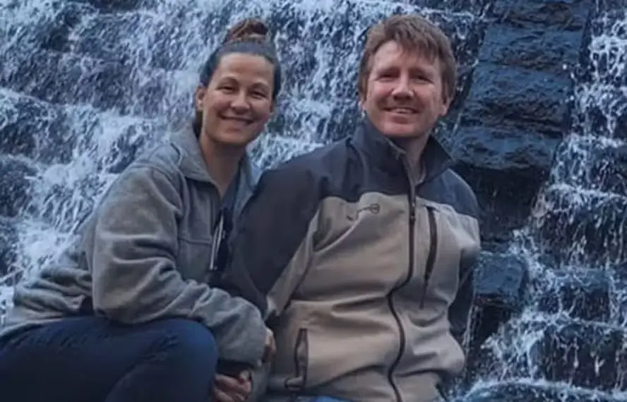 Uma mulher e um homem estão sentados bem próximos um do outro em frente a uma cascata de pedras, ambos sorrindo para a câmera e vestindo jaquetas para atividades ao ar livre.