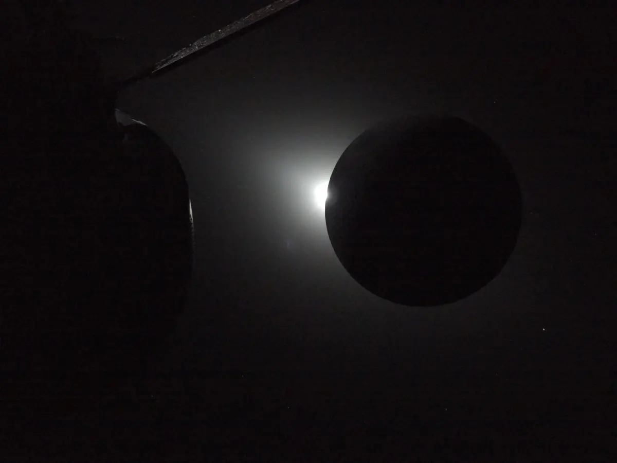 Uma grande esfera negra bloqueia parcialmente uma fonte de luz brilhante, criando um efeito de halo na escuridão ao redor. O tom é misterioso e sereno.