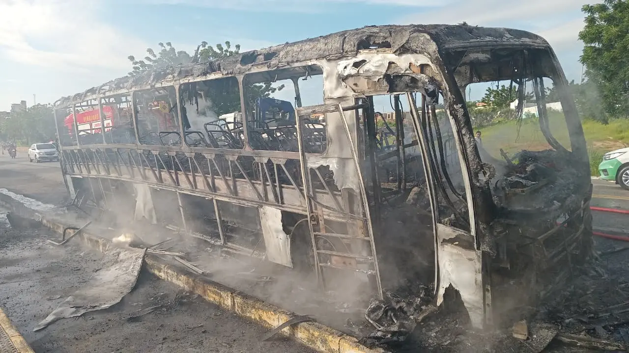 Um ônibus completamente carbonizado e em chamas repousa no canteiro central de uma rodovia, liberando uma fumaça densa e cinzenta. Ao fundo, um caminhão do Corpo de Bombeiros e outros veículos observam a cena sob um céu claro de dia.