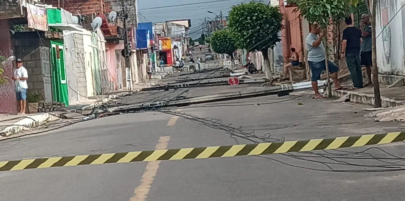Rua residencial interditada com fita de isolamento, fios elétricos espalhados pelo asfalto e postes caídos ao longo da via; moradores observam a área enquanto casas e árvores aparecem nas laterais da rua.