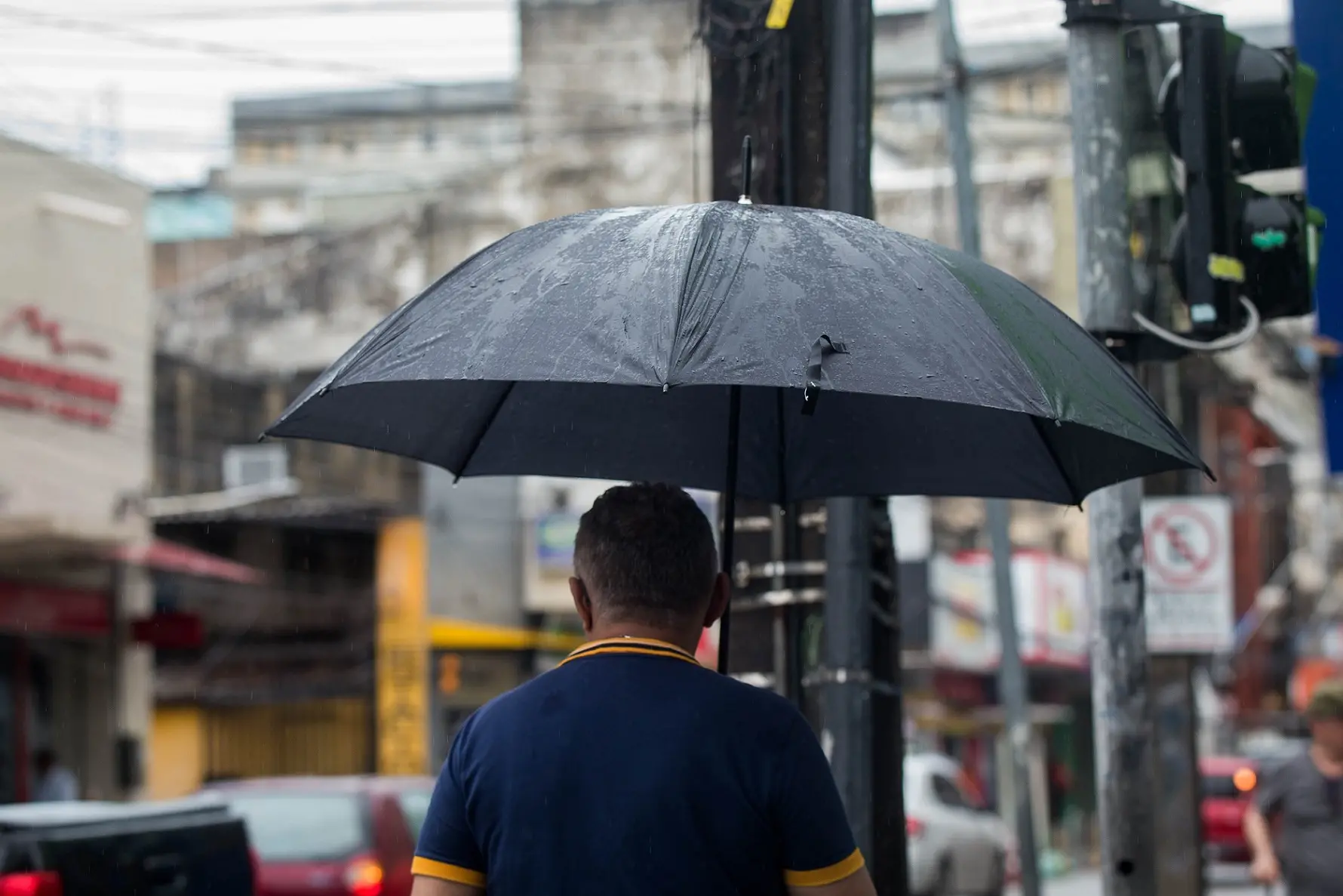 Homem de costas e blusa gola polo segurando guarda-chuva preto em dia chuvoso no Centro de Fortaleza.