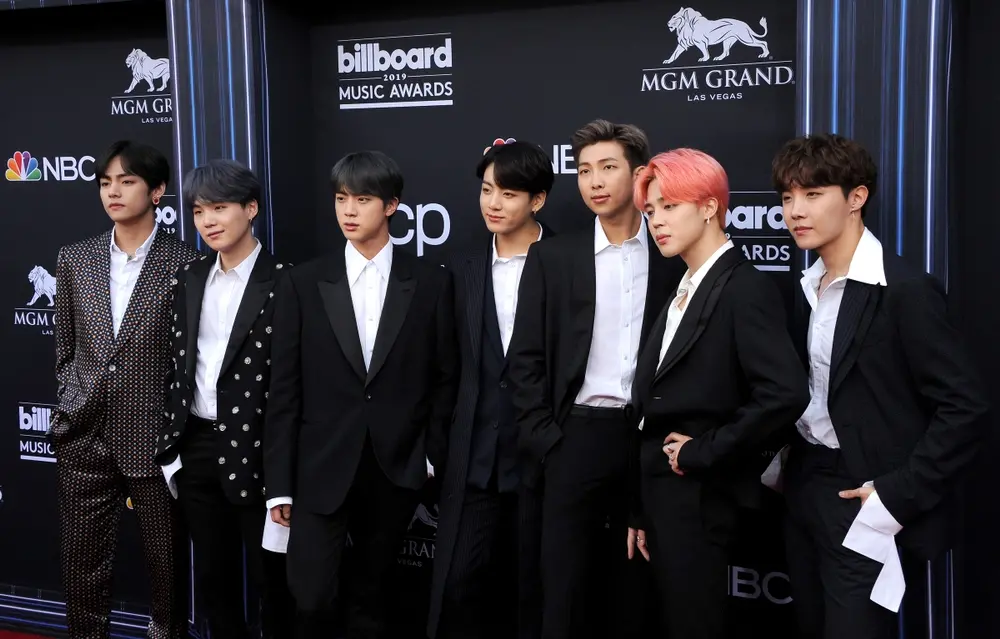 Sete homens em elegantes ternos pretos posam no tapete vermelho do Billboard Music Awards de 2019. O fundo exibe o logotipo da Billboard e placas do MGM Grand. A atmosfera é glamorosa e formal.