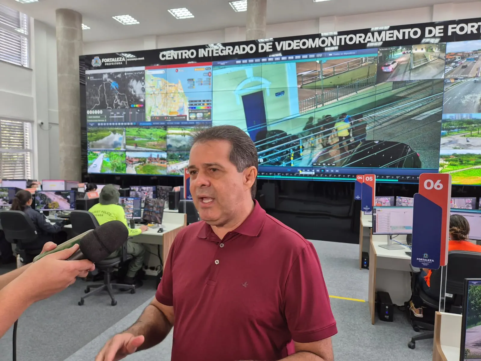 Evandro Leitão concede entrevista em centro de videomonitoramento, com microfone apontado para ele, enquanto ao fundo há um grande painel com múltiplas telas exibindo imagens de câmeras de segurança e operadores trabalhando em computadores.