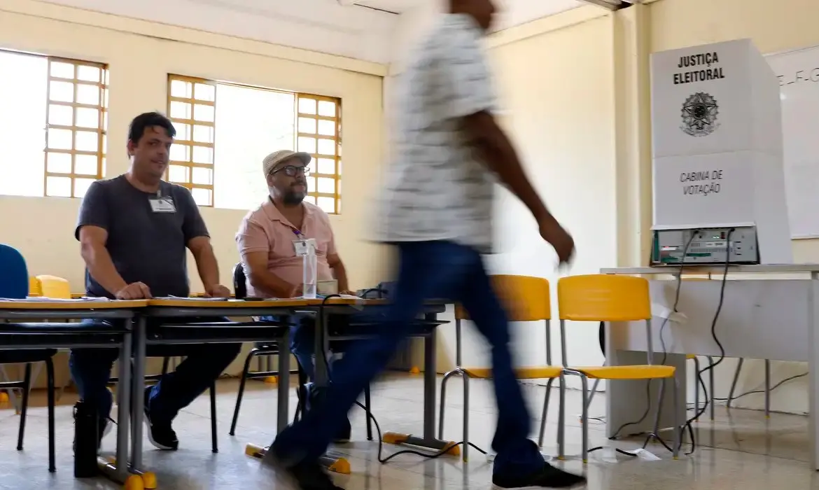 Eleitor caminha em frente a mesários em uma sala de votação, enquanto ao lado está uma cabine da Justiça Eleitoral com urna eletrônica pronta para uso.
