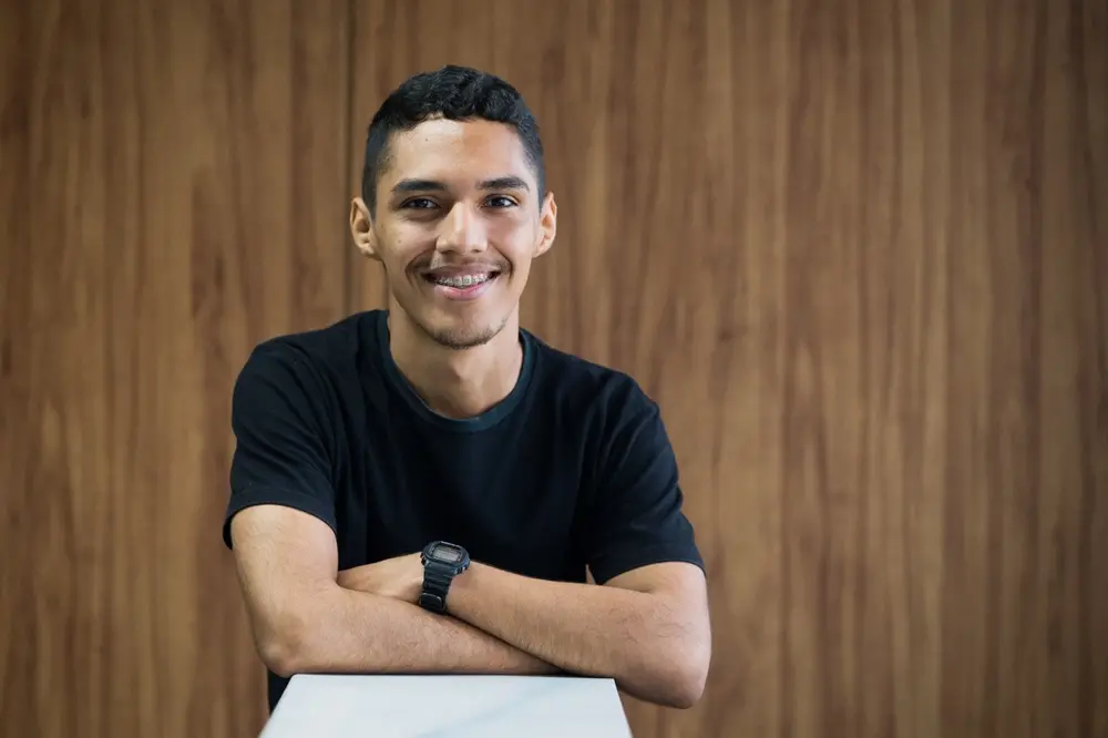Na imagem, um homem jovem de pele parda e cabelos curtos e escuros sorri para a câmera. Ele usa aparelho ortodôntico, uma camiseta preta básica e um relógio digital de pulso preto. Ele está com os braços cruzados, apoiados sobre uma superfície branca em primeiro plano. Ao fundo, há uma parede revestida de madeira com veios verticais em tons de marrom. A iluminação é suave, destacando o rosto do rapaz.