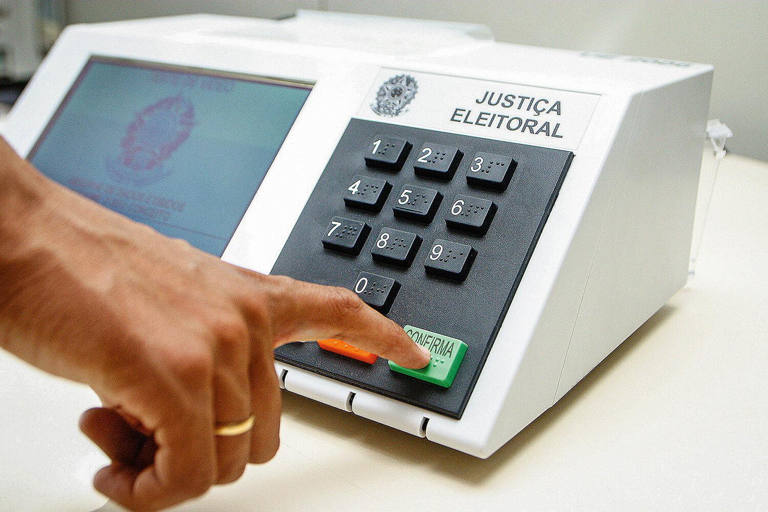 Pessoa pressiona o botão verde “CONFIRMA” de uma urna eletrônica da Justiça Eleitoral brasileira, com teclado numérico visível e tela ao lado, durante o processo de votação.