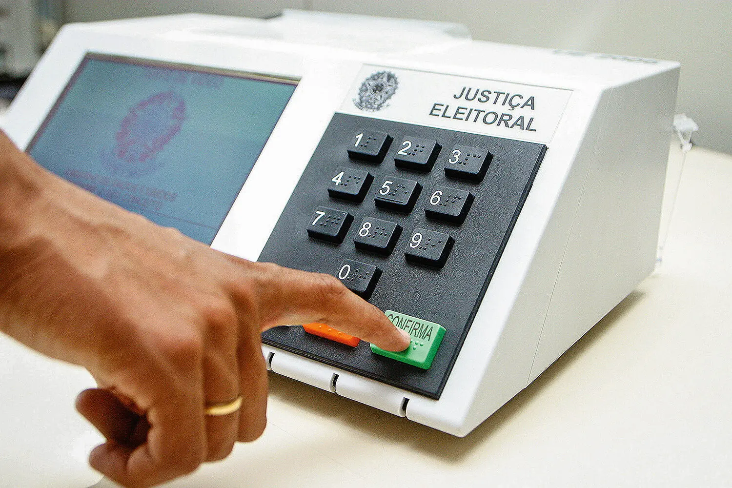 Pessoa pressiona o botão verde “CONFIRMA” de uma urna eletrônica da Justiça Eleitoral brasileira, com teclado numérico visível e tela ao lado, durante o processo de votação.