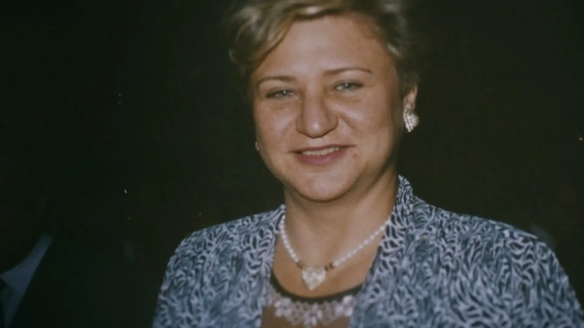 Retrato de Anita Harley, uma mulher de cabelos curtos e claros, sorrindo suavemente em um ambiente com fundo escuro. Ela utiliza um traje com estampa abstrata em tons de azul e branco, complementado por um colar de pérolas e brincos discretos.