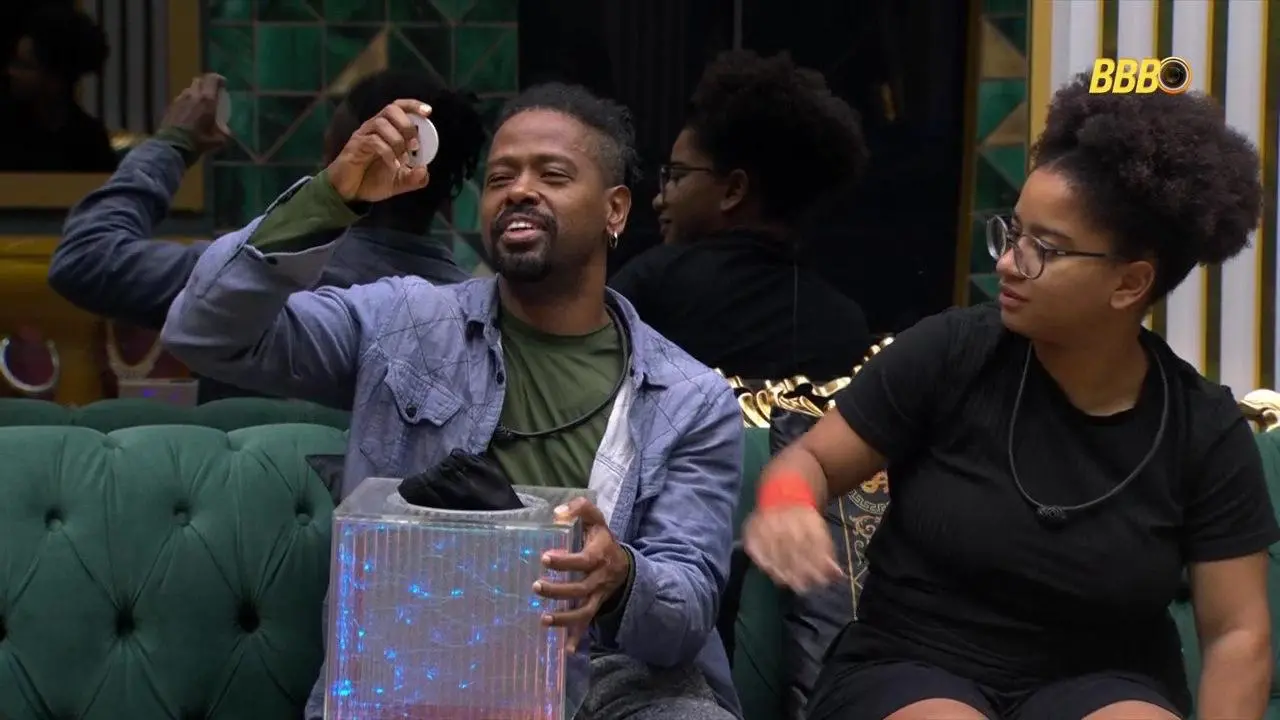 Duas pessoas sentadas em um sofá verde participam de uma dinâmica em um estúdio; uma delas segura um pequeno objeto retirado de uma caixa transparente iluminada, enquanto a outra observa ao lado.