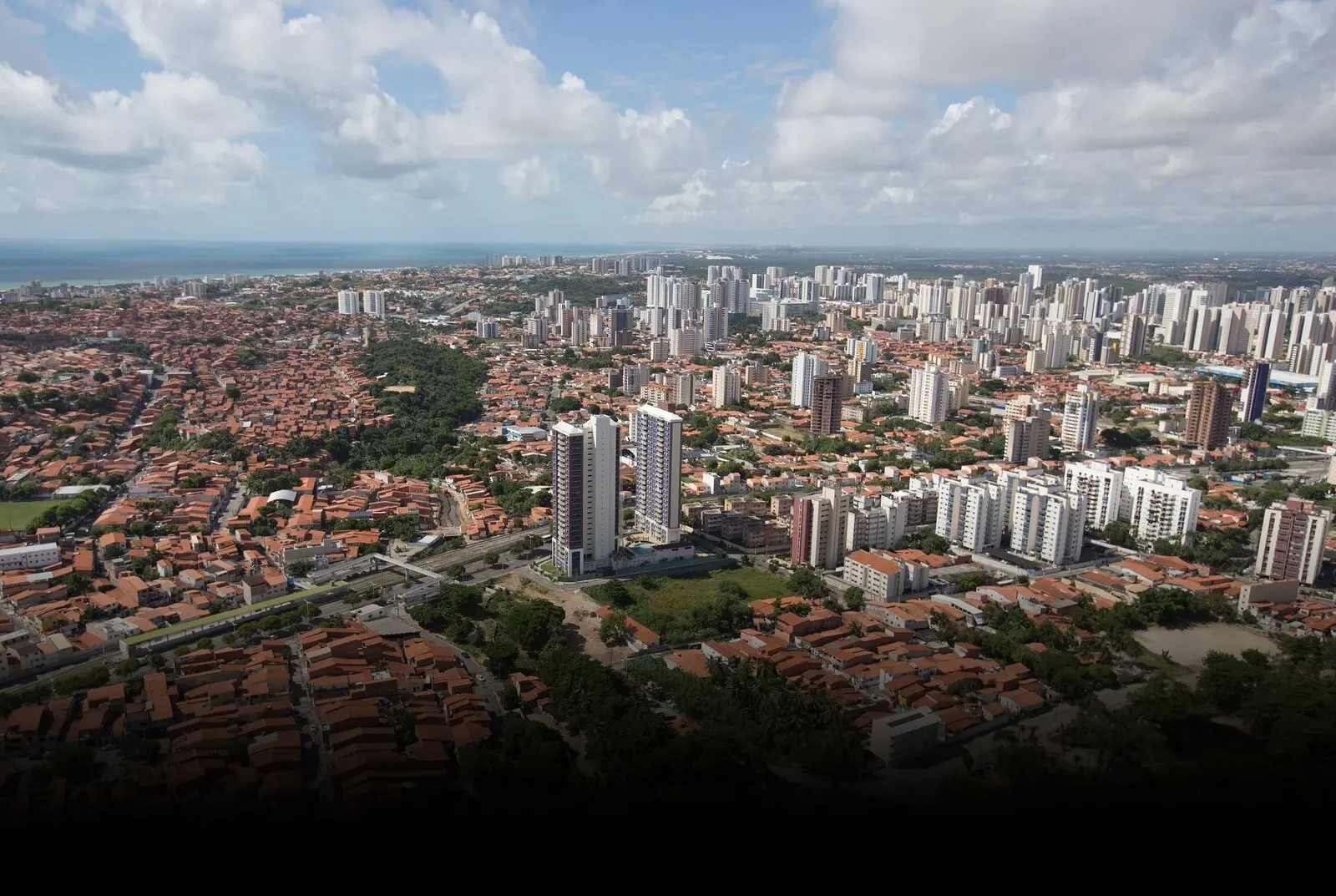 Vista aérea mostrando o contraste urbano de Fortaleza, com bairros densamente povoados de casas com telhados vermelhos em primeiro plano e grandes prédios modernos ao fundo.