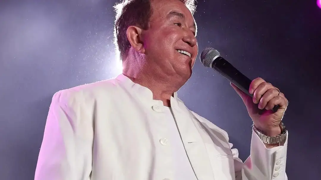 Amado Batista é um homem idoso, branco e que usa camisa branca e terninho branco. Ele está cantando segurando um microfone.