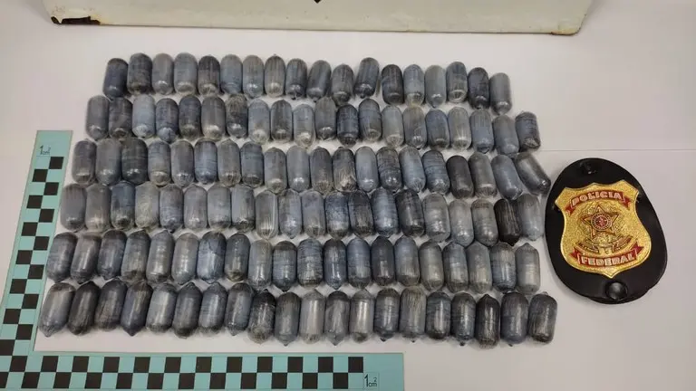 Várias cápsulas de cocaína que foram apreendidas com passageiros que tentavam embarcar de Fortaleza para Paris. Ao lado, há um distintivo da Polícia Federal.
