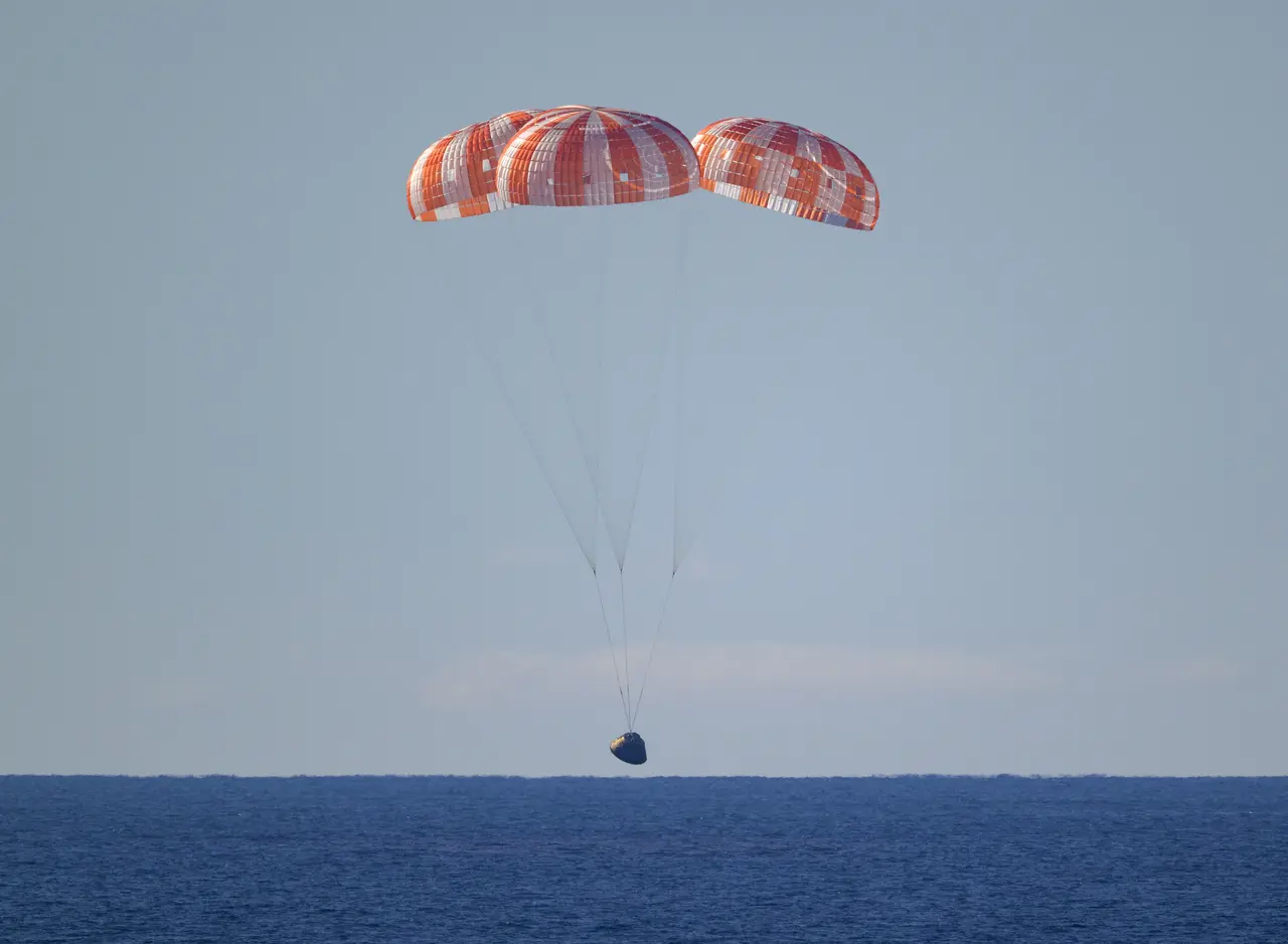 A cápsula da missão Artemis II descende suavemente em direção ao oceano, sustentada por três grandes paraquedas laranjas e brancos totalmente inflados contra o céu claro. A imagem captura o momento do 