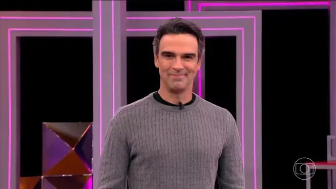 O apresentador Tadeu Schmidt sorri para a câmera no palco do Big Brother Brasil, vestindo uma blusa de tricô cinza sobre uma gola alta preta. Ao fundo, painéis geométricos com iluminação em neon rosa compõem o cenário moderno e vibrante do programa.