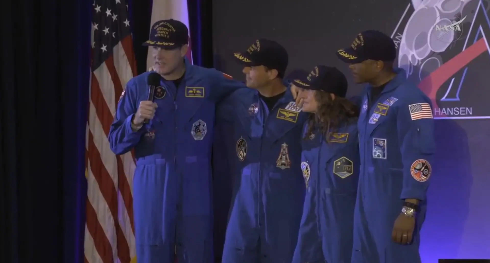 Os quatro astronautas da missão Artemis II aparecem abraçados em um palco durante uma entrevista, vestindo macacões azuis com diversos patches e bonés escuros. À esquerda, um deles fala ao microfone diante da bandeira dos Estados Unidos, em uma composição que transmite união e celebração pelo sucesso da missão.