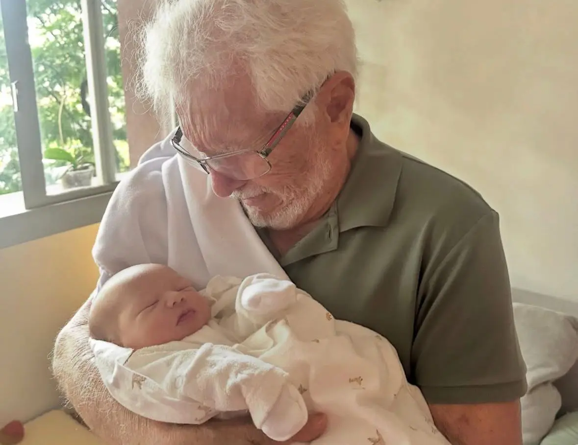 Carlos Alberto, um homem branco, idoso e de cabelos brancos, segura nos braços um recém-nascido. Eles estão em um quarto de bebê.