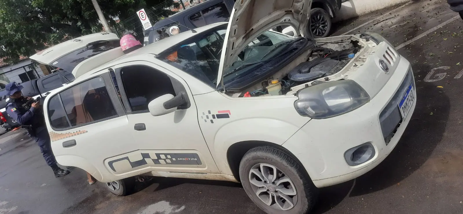 Carro branco estacionado na rua com o capô aberto, mostrando o motor; o veículo tem adesivos pretos nas portas e rodas cinza. Ao fundo, há outros carros estacionados, árvores e uma pessoa de uniforme escuro próxima à lateral traseira do automóvel, em um ambiente urbano durante o dia. Carro havia sido roubado e foi recuperado em Sobral, no Ceará.