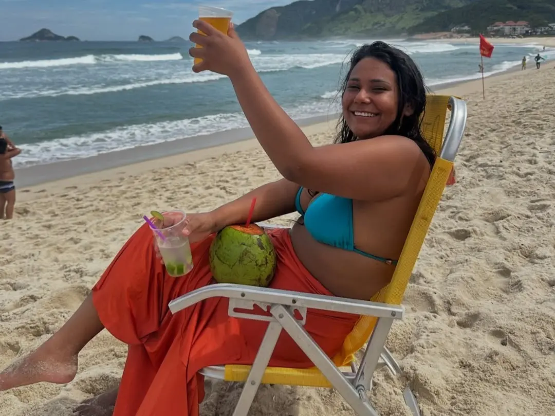 Foto que contém Chaiany Andrade, participante do BBB 26. Ela veste um biquíni azul, uma saia laranja, segura na mão esquerda um copo de cerveja, na direita uma caipirinha e no colo um coco verde. Ela está sentada em uma cadeira amarela de praia, em faixa de areia.