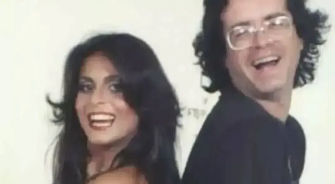 Duas pessoas posicionadas de costas uma para a outra, em enquadramento próximo, usando roupas escuras sobre fundo claro.