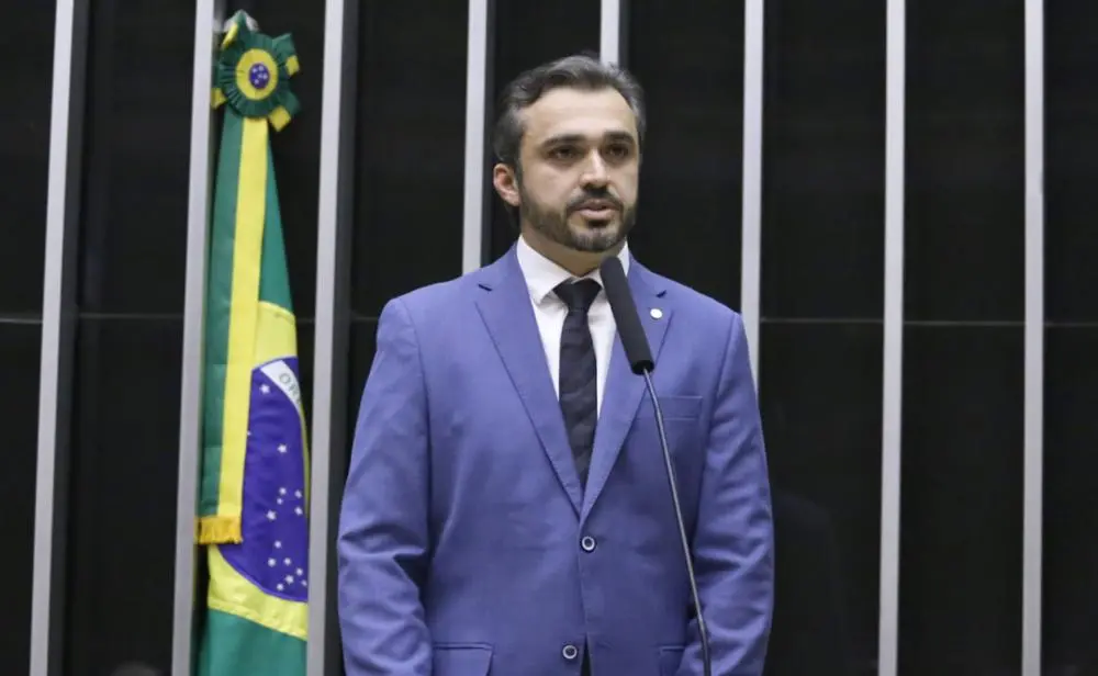 Homem de terno azul fala ao microfone em tribuna, com a bandeira do Brasil ao fundo, em ambiente que remete a uma sessão legislativa.