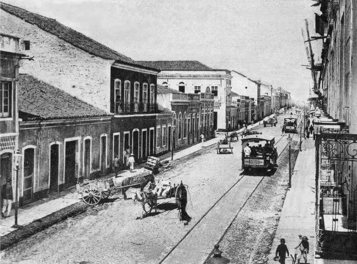 Registro histórico em preto e branco da Rua Barão do Rio Branco no início do século XX, mostrando trilhos de bonde, carroças de tração animal e casarões coloniais preservados.