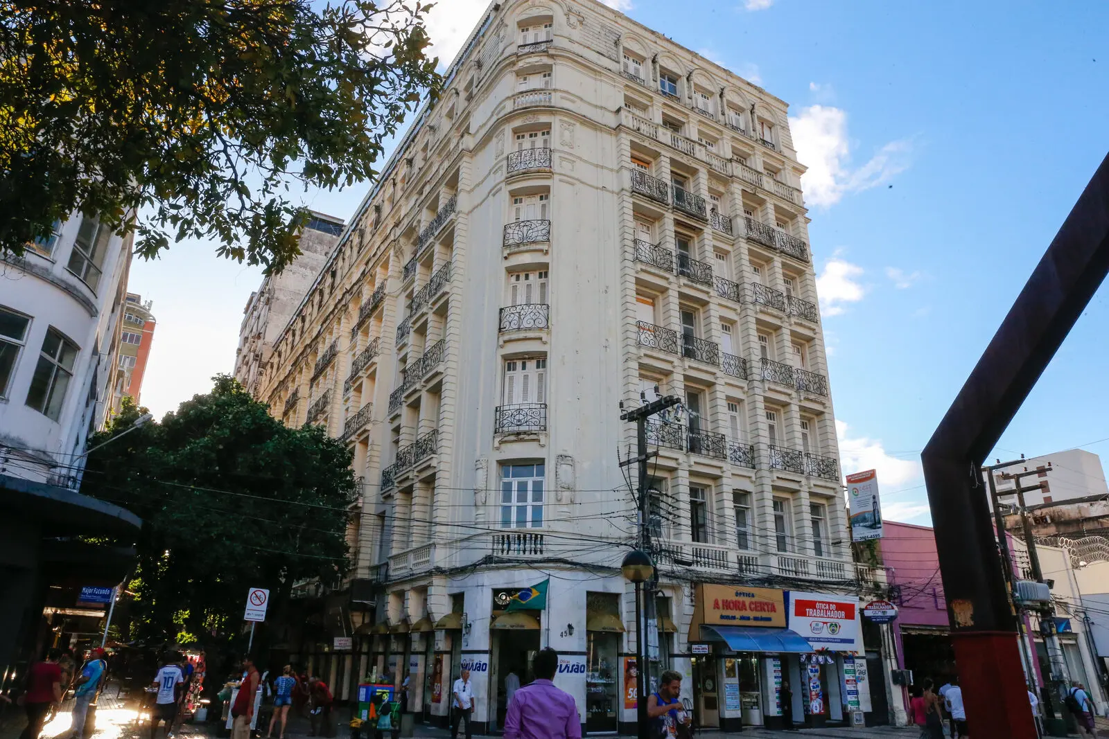 Fachada do histórico Edifício Excelsior, no Centro de Fortaleza, exibindo sua arquitetura clássica com várias janelas e sacadas de ferro, situado em uma esquina movimentada com comércio local.