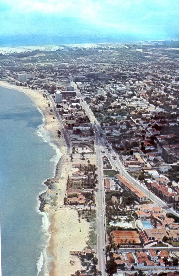 Imagem aérea da orla de Fortaleza nos anos 70, exibindo a Avenida Beira-Mar com poucos edifícios altos, amplas áreas verdes, clubes de lazer e as dunas ao fundo.
