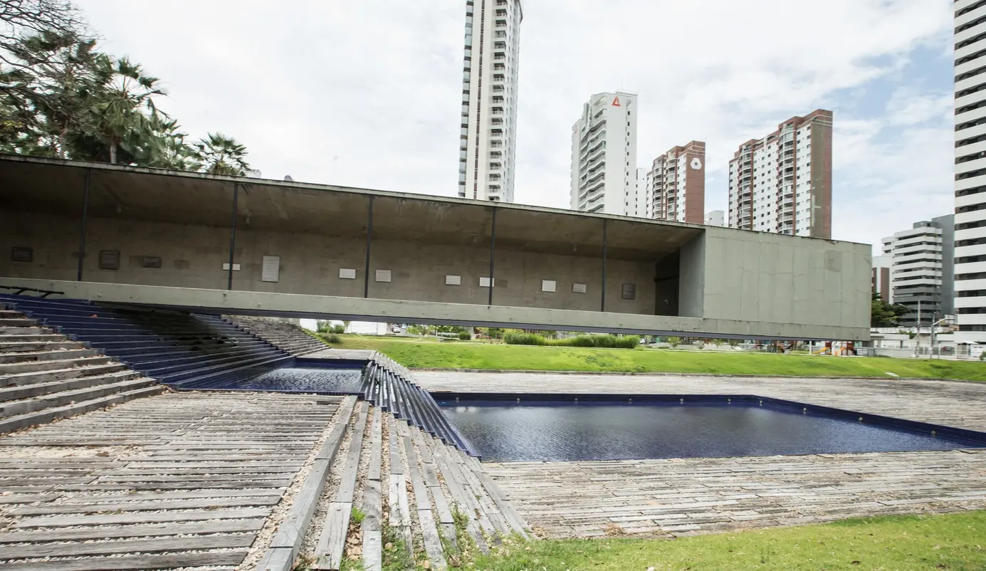 Mausoléu Castelo Branco: estrutura brutalista com uma monumental caixa de concreto aparente em balanço sobre um espelho d'água azul. À esquerda, uma rampa de madeira e cascalho conduz ao monumento, cercado por gramado e árvores. Ao fundo, prédios modernos da Aldeota contrastam com a horizontalidade radical da obra de Sérgio Bernardes.