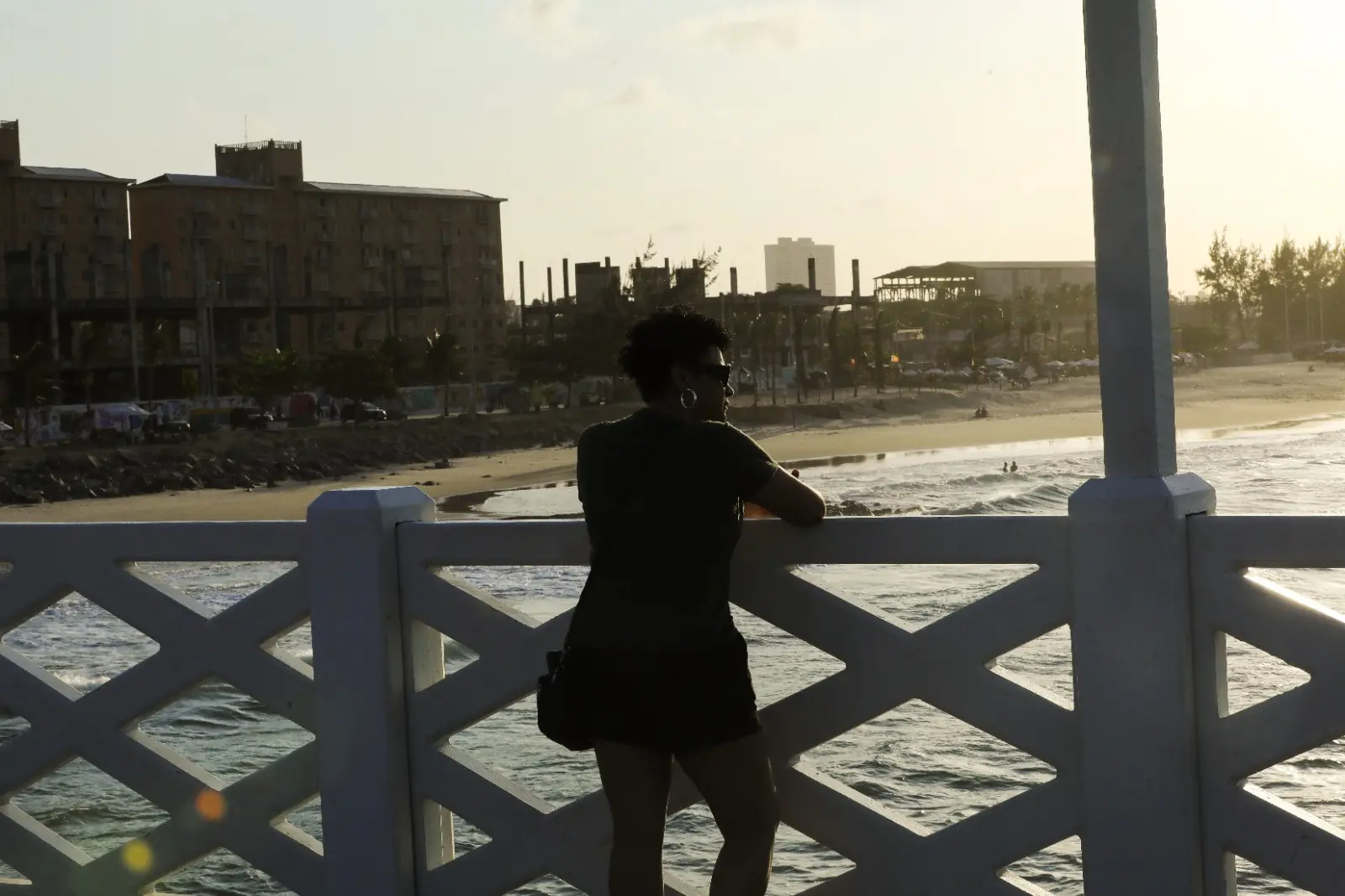 Pessoa apoiada em um parapeito branco observa o mar ao entardecer. Ao fundo, há uma praia com algumas pessoas, prédios e estruturas urbanas iluminadas pela luz dourada do sol baixo.