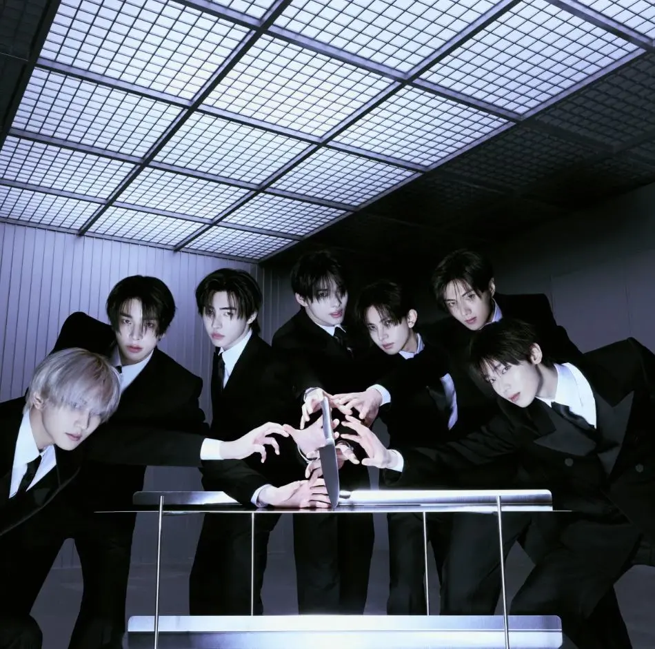 Grupo de pessoas vestidas com ternos pretos estende as mãos em direção a um objeto central sobre uma mesa metálica, em ambiente interno com iluminação fria e teto quadriculado.