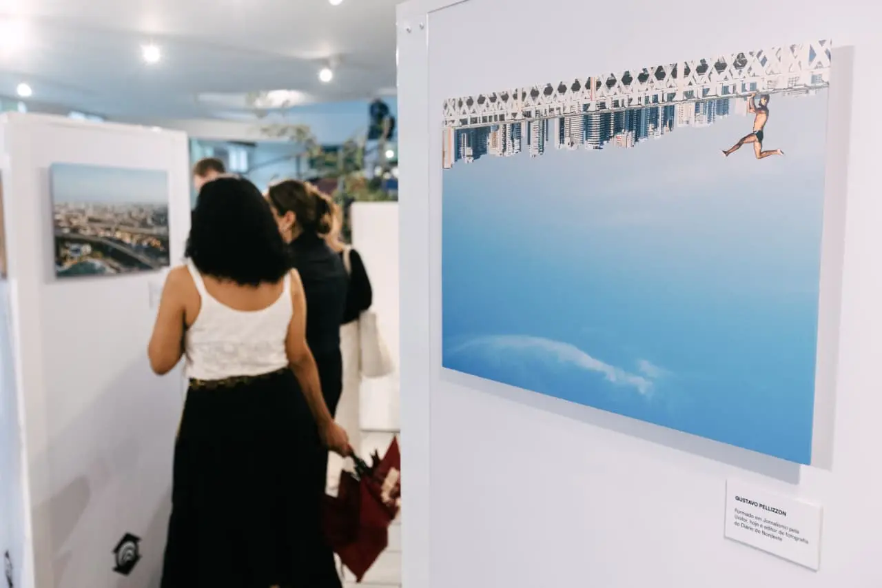 Na imagem, fotografia em plano médio de uma galeria de arte ou exposição fotográfica. Em primeiro plano, à direita, destaca-se uma obra retangular fixada em uma parede branca. A fotografia na obra apresenta uma composição surrealista: a parte superior mostra uma cidade costeira e uma ponte invertidas (de cabeça para baixo), com um homem saltando no ar em direção ao que seria o céu, mas que ocupa a maior parte inferior da imagem em um tom azul límpido com poucas nuvens. Abaixo do quadro, há uma pequena etiqueta de identificação com o nome Gustavo Pellizzon. Ao fundo, à esquerda e levemente desfocadas, duas mulheres observam outras obras expostas. Uma delas está de costas, veste uma regata branca e saia preta, segurando um guarda-chuva fechado. O ambiente é bem iluminado, com paredes brancas e um teto alto, sugerindo um espaço cultural moderno.