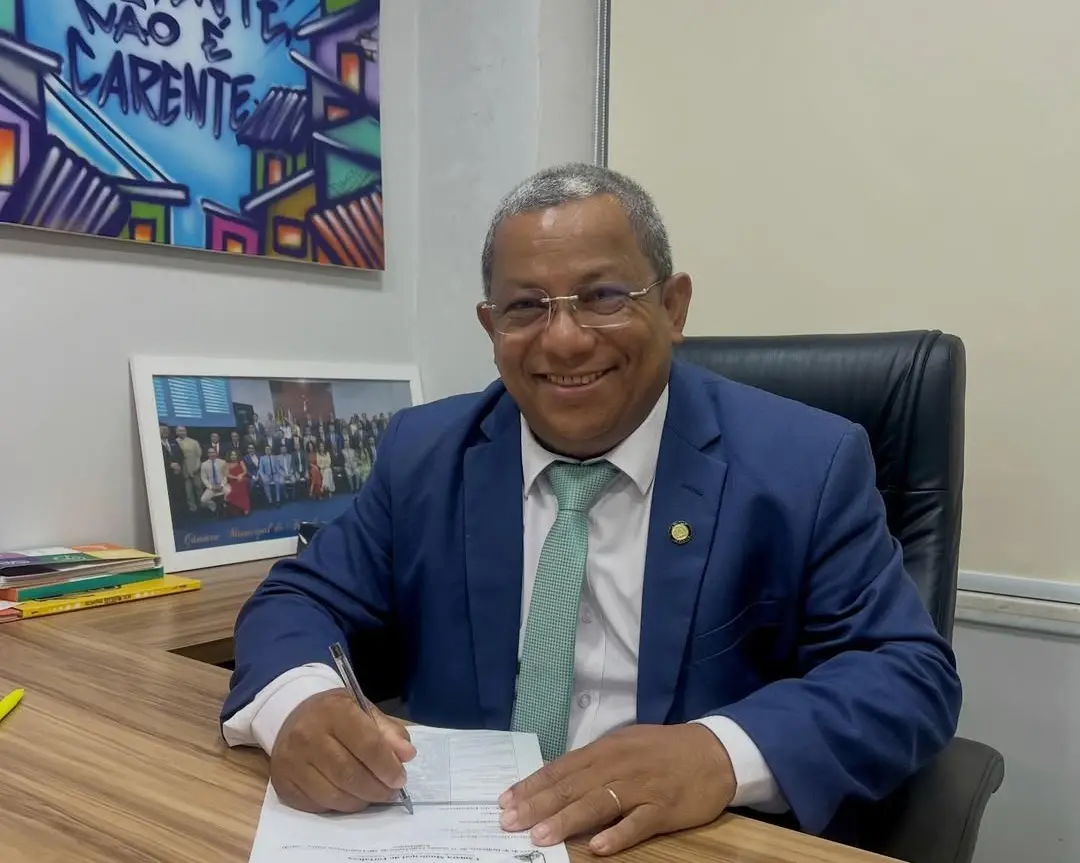 Homem de meia-idade, de pele morena, cabelo curto grisalho e óculos, sentado em uma cadeira de escritório atrás de uma mesa de madeira. Ele sorri enquanto escreve em um papel com uma caneta. Veste terno azul, camisa branca e gravata verde. Ao fundo, há um quadro colorido na parede e uma foto emoldurada com um grupo de pessoas sobre a mesa.