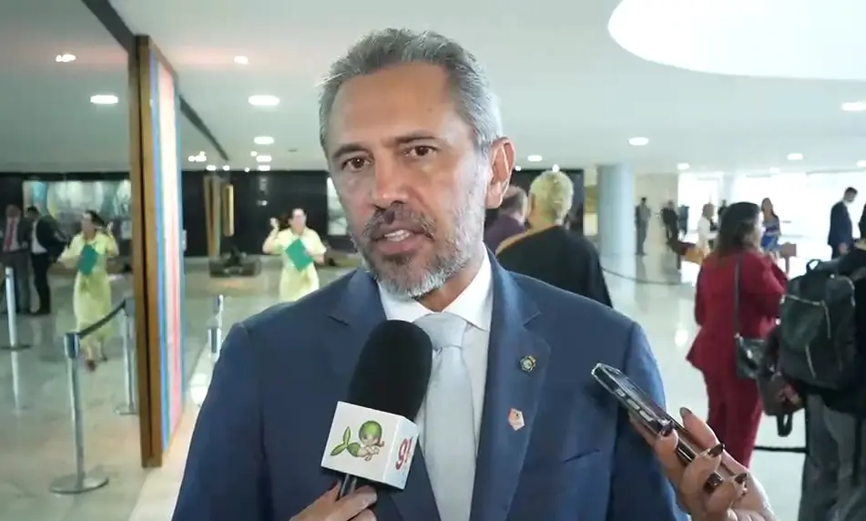 Homem de meia-idade, com cabelo grisalho curto e barba aparada, veste terno azul, camisa branca e gravata clara. Ele está sendo entrevistado em um ambiente interno amplo e bem iluminado, possivelmente um prédio institucional, enquanto fala diante de um microfone com logotipo. Ao fundo, há várias pessoas caminhando ou conversando, algumas com pastas e celulares, e o espaço possui piso brilhante e iluminação no teto.