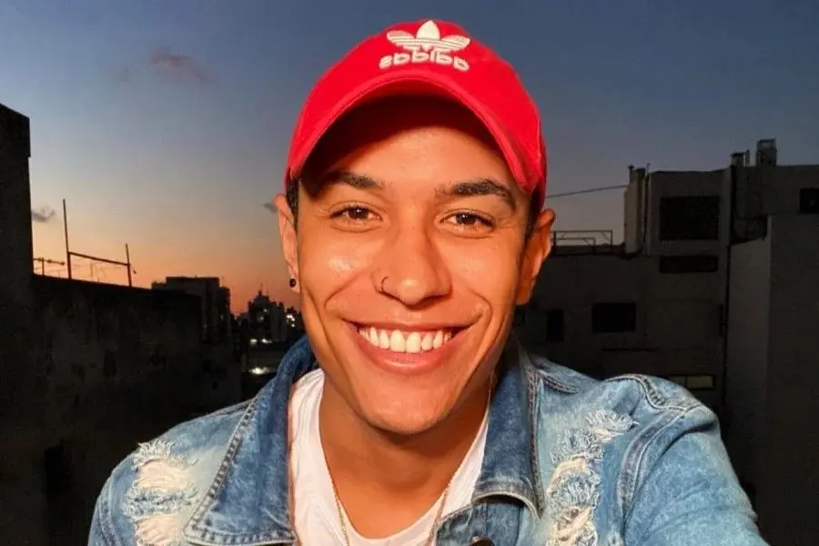 Jovem sorridente usando boné vermelho e jaqueta jeans ao pôr do sol, em ambiente urbano, capturando uma foto para redes sociais.