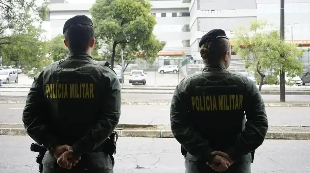 Dois policiais da Polícia Militar do Ceará vistos de costas, com as mãos para trás, em área externa urbana; prédios, árvores e veículos aparecem ao fundo.
