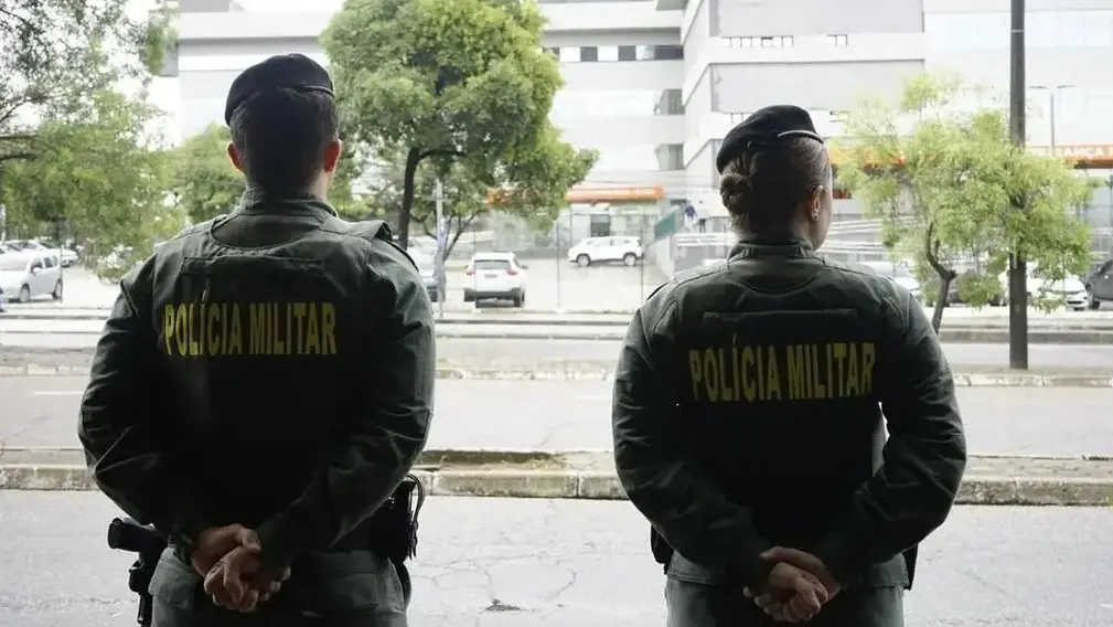 Dois policiais da Polícia Militar do Ceará vistos de costas, com as mãos para trás, em área externa urbana; prédios, árvores e veículos aparecem ao fundo.