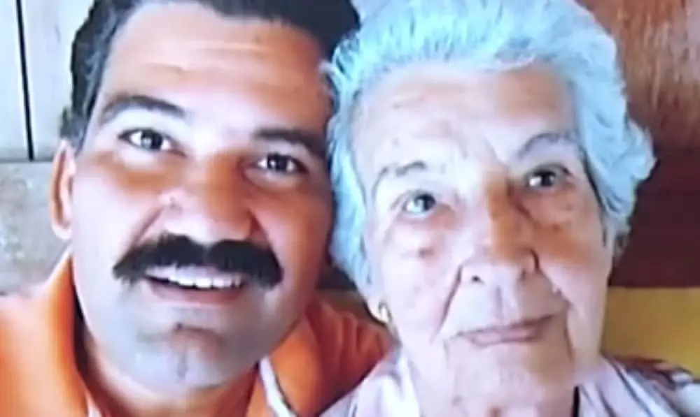 Um homem de bigode sorri ao lado de uma senhora idosa de cabelos brancos em um registro próximo e afetuoso. A imagem captura o contraste entre as expressões dos dois em uma composição simples e iluminada.