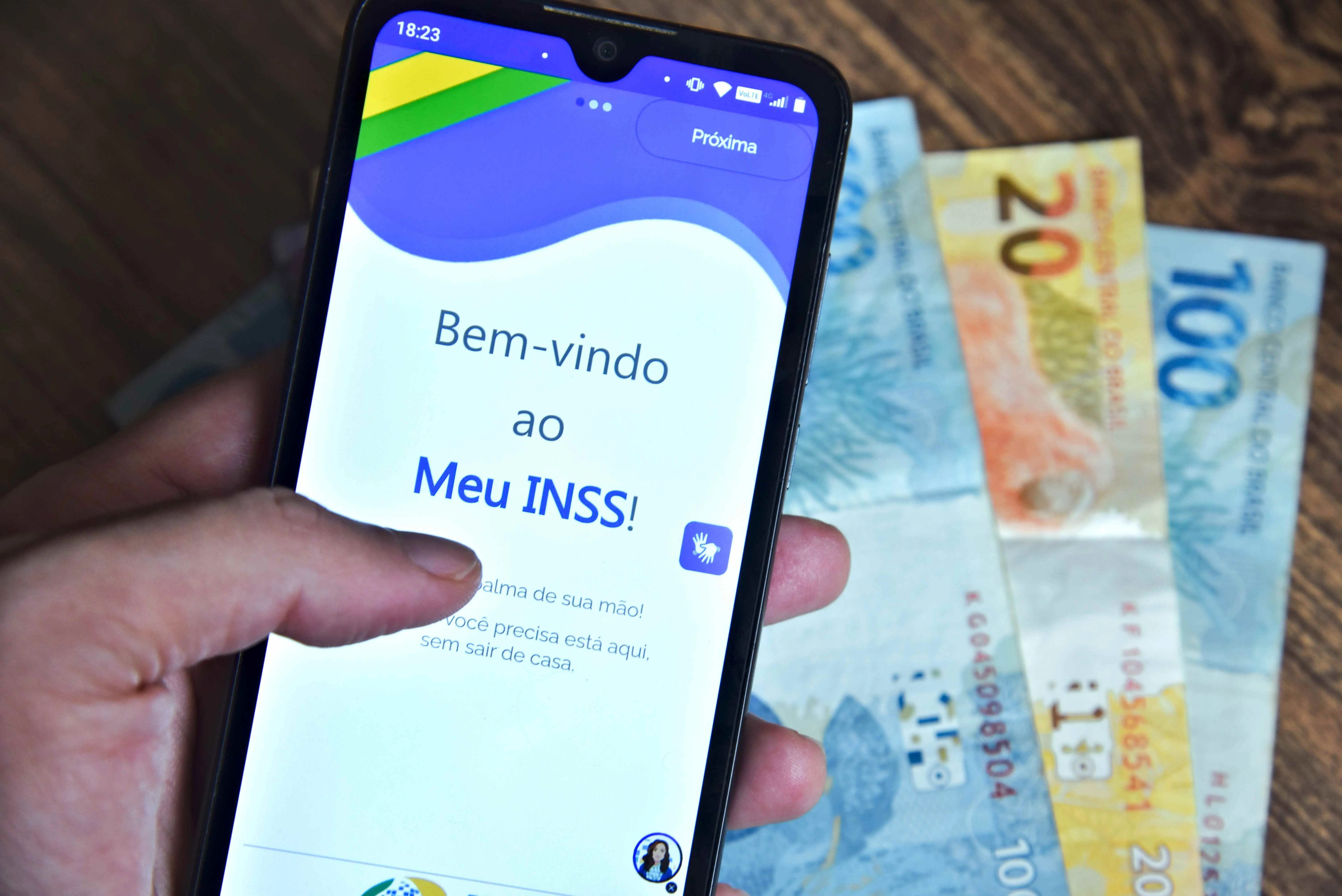 Pessoa segurando celular, com aplicativo do Meu INSS na tela, e cédulas de dinheiro ao fundo.