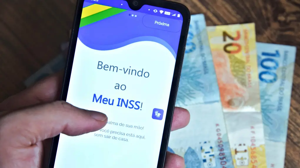 Pessoa segurando celular, com aplicativo do Meu INSS na tela, e cédulas de dinheiro ao fundo.