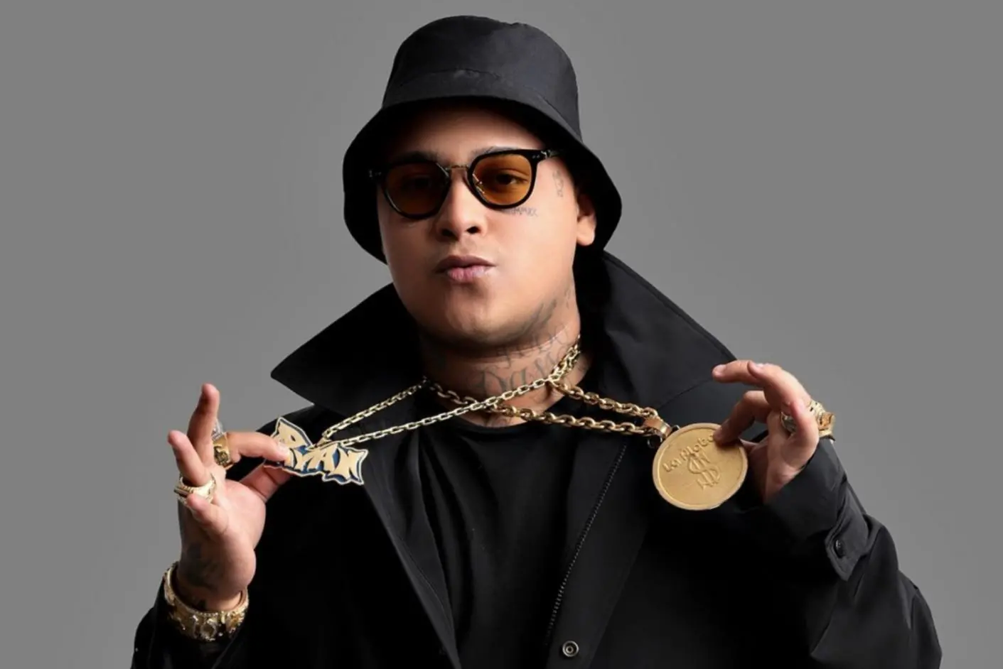 Pessoa vestindo roupas pretas posa diante de fundo cinza, usando chapéu bucket e óculos escuros, e segura com as duas mãos um colar de corrente dourada com um pingente grande e outro em formato de letra estilizada.