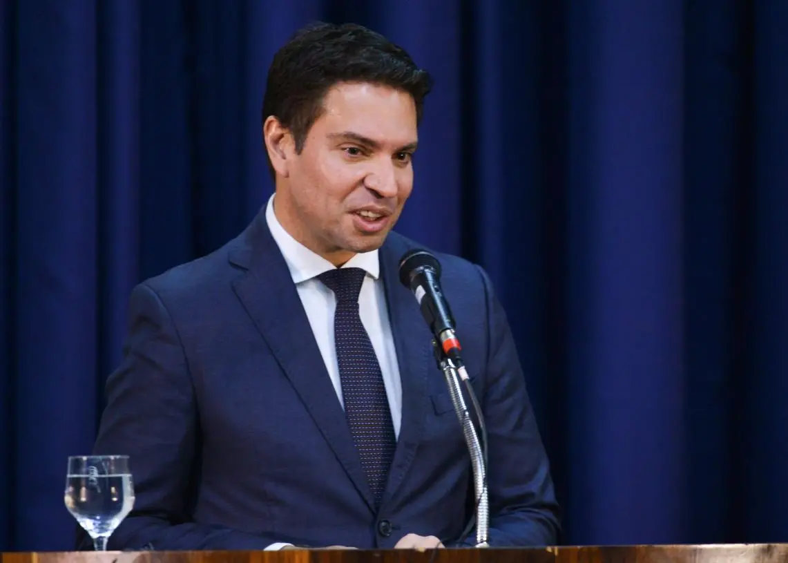 Pessoa de terno escuro fala ao microfone atrás de um púlpito de madeira, com cortina azul ao fundo; um copo com água está à esquerda.