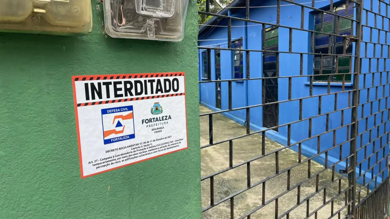 Um adesivo da Defesa Civil de Fortaleza com o aviso 'INTERDITADO' está colado em uma parede verde na entrada de uma vila, próximo a um medidor de energia. Ao fundo, uma grade de ferro preta delimita o acesso a uma casa azul, em uma composição que evidencia o isolamento da área por questões de segurança.